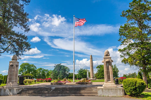 An American flag flies in the wind at the top of stairs leading to the Walnut Hill Park rose garden in New Britain, Connecticut.  The city's World War I memorial is in the background to the right of the flagpole.