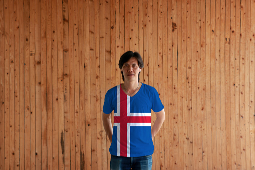 Man wearing Iceland flag color of shirt and standing with crossed behind the back hands on the wooden wall background, blue sky with a snow-white cross and a fiery-red cross inside the white.