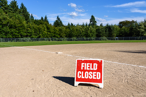 Empty local baseball field on a sunny day with woods and blue sky in the background, Field Closed sign