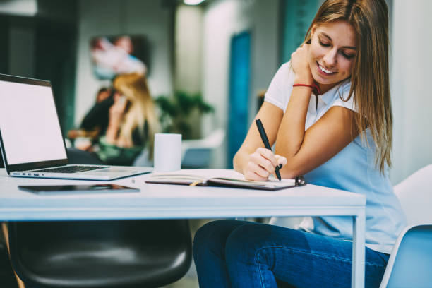 istock Cheerful young woman writing down text information in notepad doing homework and sitting at desktop with modern laptop computer with blank screen area for your internet content or website in office 1159338324