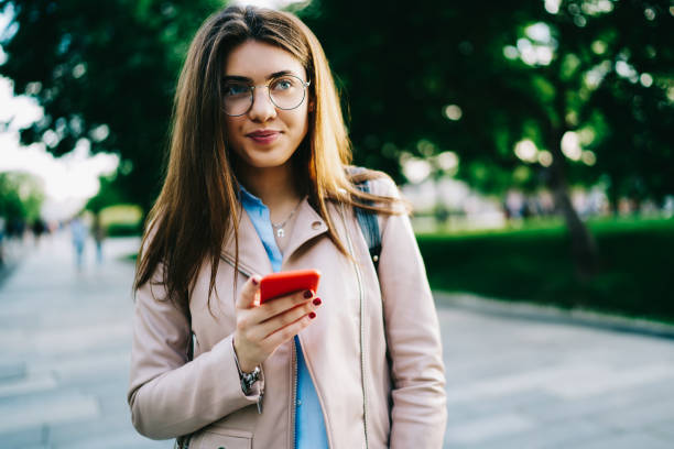 istock Pensive young woman in eyewear thinking about making online booking holding smartphone while standing on street, attractive hipster girl connecting mobile phone to 4G internet resting on spring day 1159339442