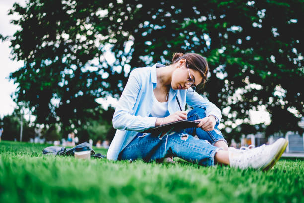 istock Thoughtful female student in eyewear making homework task planning project during rest in green park, pensive hipster girl concentrated on studying drawing sketches sitting on grass on leisure 1159339664