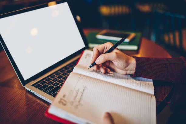 Cropped image of female's copywriter hands writing down information to notepad while sitting at wooden table and near to laptop with blank copy space screen for your advertising information content imagen recortada de las manos de la redactora femenina escribiendo información en el bloc de notas mientras se sienta en la mesa de madera y cerca de la computadora portátil con pantalla de espacio de copia en blanco para su contenido de información pub