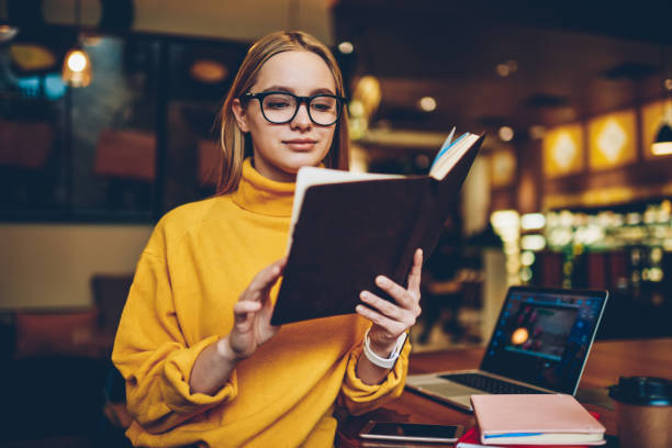 istock Young attractive positive female student in spectacles for eye correction reading interesting literature during exam preparation.Cute intelligent hipster girl analyzing information from notepad 1159341316