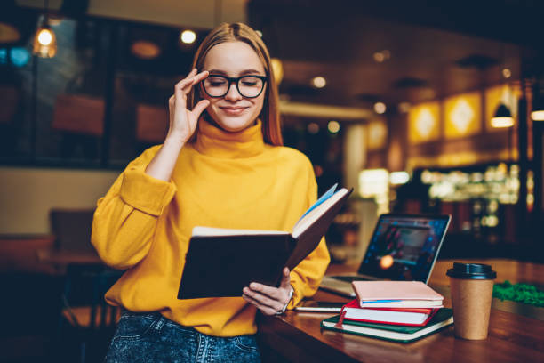 istock Prosperous female blogger correcting glasses for better eye vision while reading own publication notations from textbook.Charming happy blonde woman enjoying time indoors with interesting literature 1159341323