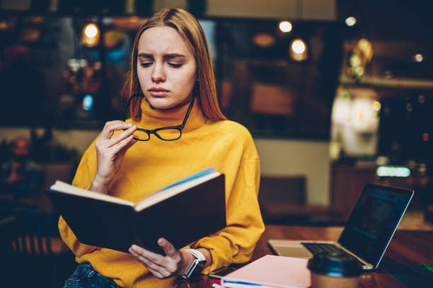 istock Charming talented sad female student wearing cool eyeglasses for better vision while preparing for upcoming examinations indoors.Cute disappointed hipster girl reading literature in coffee shop 1159341333