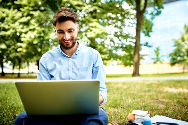 homem de negócios de sorriso novo que senta-se na grama no parque durante o almoço e que trabalha no portátil - laptop computer grass nature - fotografias e filmes do acervo