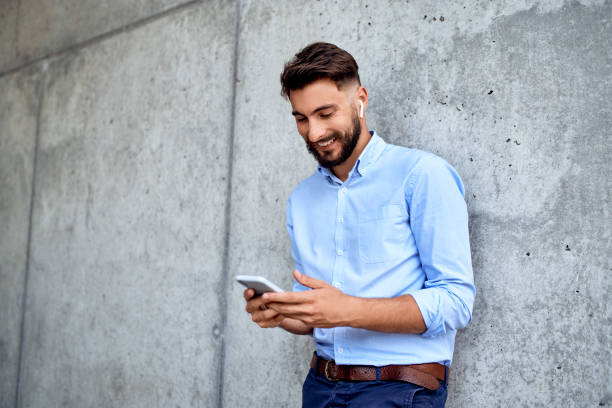 istock Cheerful young businessman wearing wireless headphones and using phone while leaning against wall 1159350349