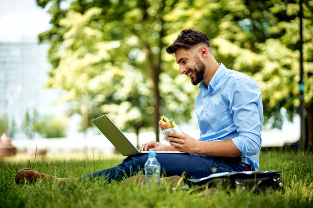 Young cheerful businessman sitting in the park with sandwich and laptop Young cheerful businessman sitting in the park with sandwich and laptop businessman happiness outdoors cheerful stock pictures, royalty-free photos & images