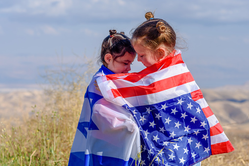 Two cute girls with American and Israel flags. Little children holding Israeli and USA flags hugging on meadow with beautiful landscape in background.