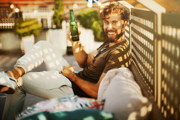 Enjoying rooftop party and summer days. Side view of young man enjoying rooftop party. He's sitting on sofa, smiling, toasting with beer and looking at camera. beer alcohol stock pictures, royalty-free photos & images