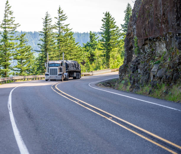 istock Classic big rig semi truck transporting covered cargo on flat bed semi trailer running on the mountain winding road 1159360404