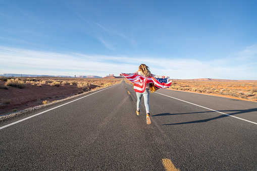 Woman running on long empty highway road leading to Monument Valley in USA holding an American flag in the air. People travel America concept