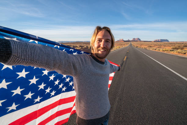 istock Man on road takes selfie while holding American flag; road trip concept 1159368418