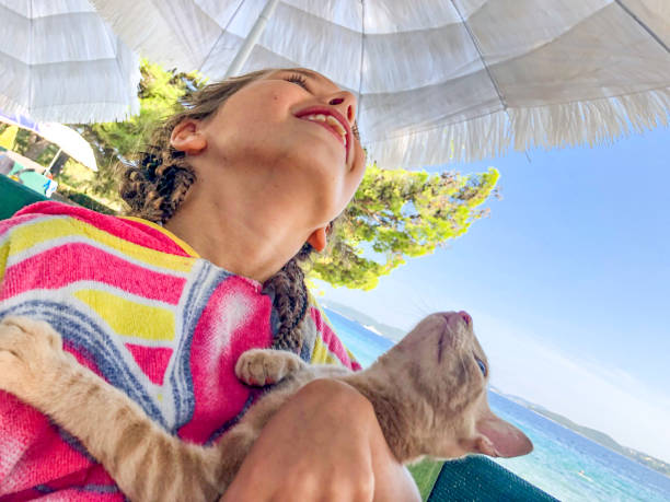 Little smiling girl petting a ginger cat sitting on a stone fence by the sea. High quality photo