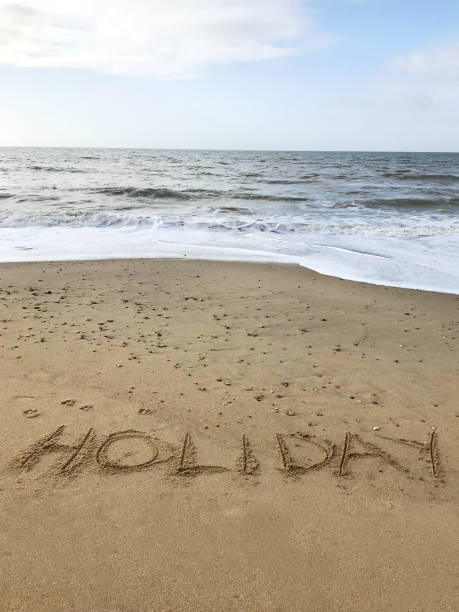 Stock photo of writing drawn on sunny beach with Holiday word written in sand with stick, by sea waves, concept social media photo of handwriting letters in soft golden sand on Bournemouth beach, Dorset, England seaside coastline view imagen de escritura dibujada en playa soleada con palabra navideña escrita en arena con palo, por las olas del mar, concepto de foto de medios sociales de letras de escritura a mano en suave arena dorada en la playa de bournemouth, dorset, inglaterra vist