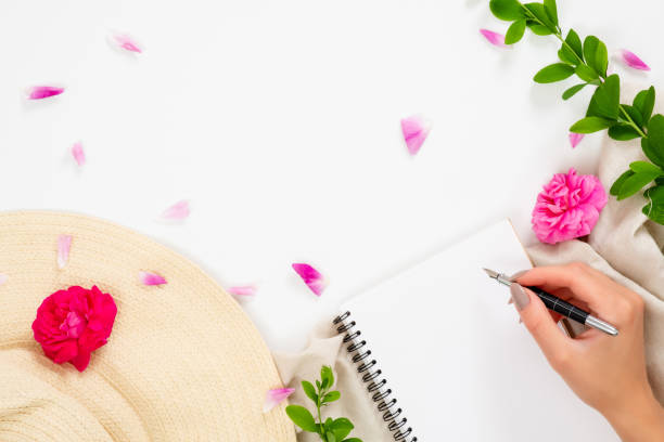 istock Flat lay, top view office table desk. Workspace with straw hat, blank paper notepad, human hand holding pen, pink rose flower buds and petal on white background. 1159373784