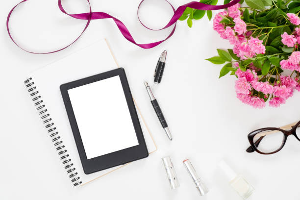 istock Modern home office desk workspace with blank screen tablet e-book, feminine accessories, glasses, paper notebook, bouquet of pink rose flowers on white background. Flat lay female table, top view. 1159373853