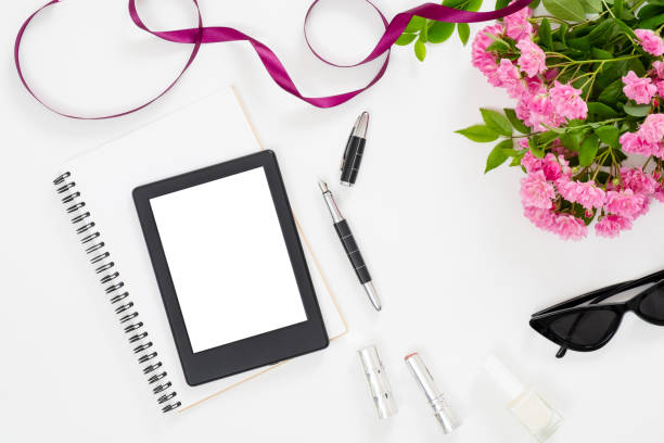 istock Modern home office desk workspace with blank screen tablet e-book, feminine accessories, glasses, paper notebook, bouquet of pink rose flowers on white background. Flat lay female table, top view. 1159373861