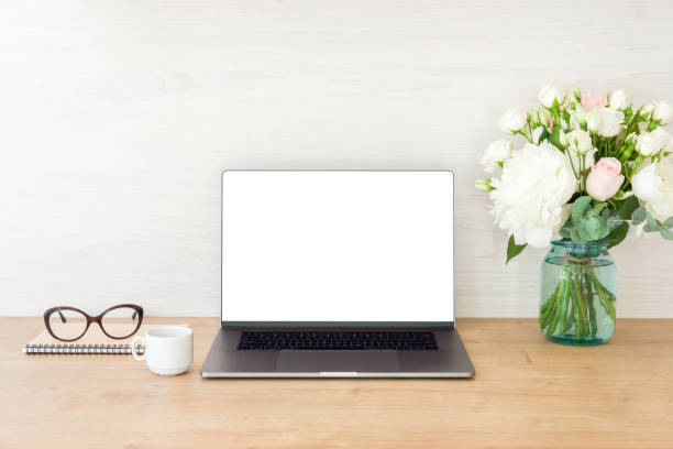 istock Office desk table with laptop computer with blank screen mockup and flowers bouquet, coffee cup, glasses, notepad on wooden surface. Minimal style feminine workplace concept. 1159375480