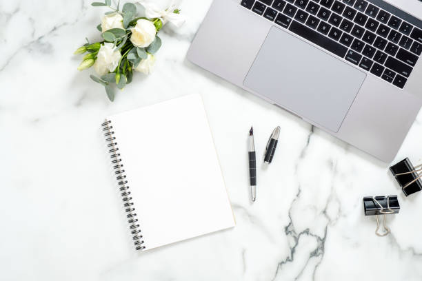 istock Flat lay home office desk. Female workspace with laptop computer, white roses flowers bouquet, accessories, diary on white marble background. Top view feminine background. 1159376149