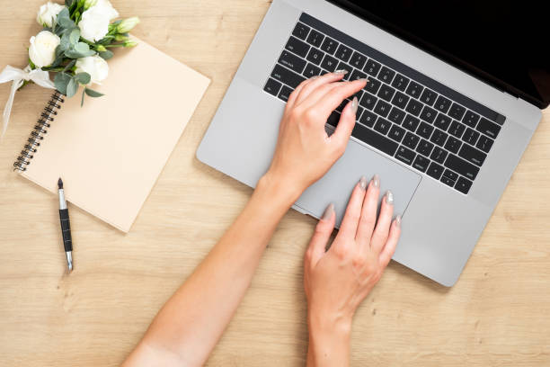 istock Wood office desk table with laptop computer, female hands typing on keyboard, paper notebook, bouquet of flowers. Stylish feminine workspace. Flat lay composition, top view, overhead. 1159376180
