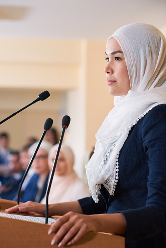 Young pretty confident muslim female delegate in hijab standing by tribune while making speech for foreign partners or colleagues