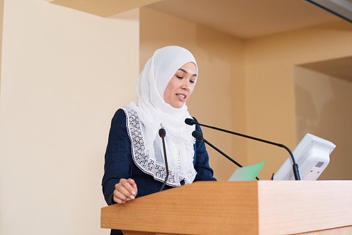 Young confident muslim businesswoman in suit and hijab reading points of her report by tribune in front of audience