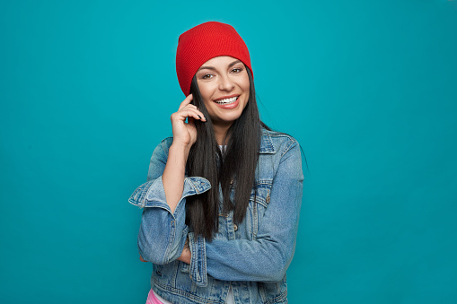Pretty, positive model in stylish clothes posing on blue background, smiling. Cheerful girl wearing in stylish red knitted hat, denim jacket. Concept of street fashion.