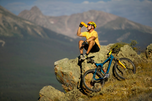Man on Rock Drinking From Water Bottle with Bicycle and Mountain View