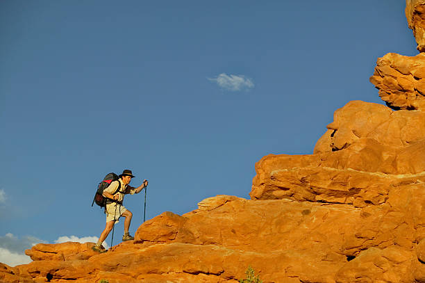 Man Hiking Up Rock Ledge Man with Backpack and Hat Hiking Up Rock Ledge in Canyonlands of Utah climbing up a hill stock pictures, royalty-free photos & images