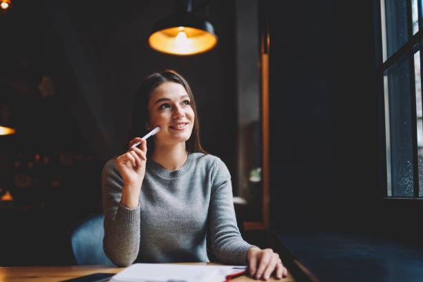 istock Dreamy hipster girl with pen in hand thinking on creative ideas for writing interesting article sitting at table with notepad in coworking.Thoughtful smiling student pondering on plans on future 1159396180