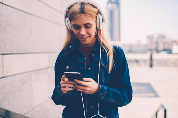 istock Smiling hipster girl in denim jacket reading incoming notification on smartphone while listening audio songs in modern headphones walking in urban setting.Cheerful student chatting online on telephone 1159398818