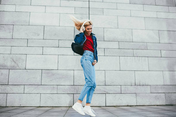 a mulher nova loura positiva vestiu-se no fato da sarja de nimes que salta durante a música elecronic fresca no fundo relativo à promoção cinzento. menina alegre do hipster que escuta canções audio favoritas em auscultadores modernos - elecronic - fotografias e filmes do acervo