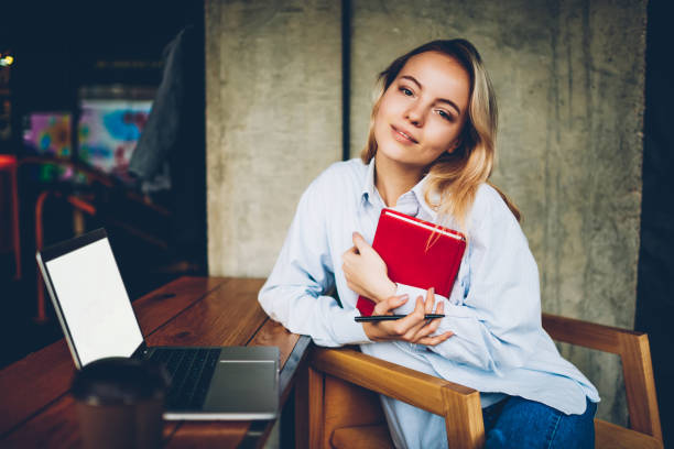 istock Portrait of charming blonde hipster student with notepad in hands liiking at camera while sitting at digital netbook with blank screen area in coworking space.Gorgeous young woman with textbook 1159399996