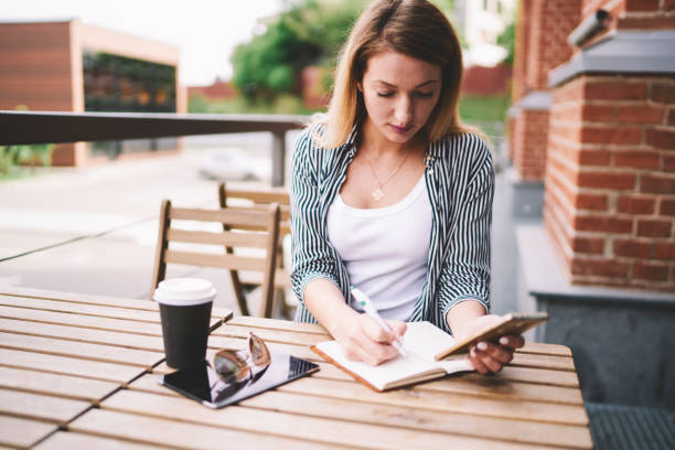 istock Concentrated female student studying at street cafeteria writing essay in notepad while using mobile phone, serious hipster girl research information for project via new modern smartphone outdoors 1159400093