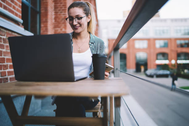 istock Young graphic designer watching online comedy show using new modern laptop while sitting at comfortable street cafe, happy hipster girl in eyeglasses share content at profile in social network 1159400127