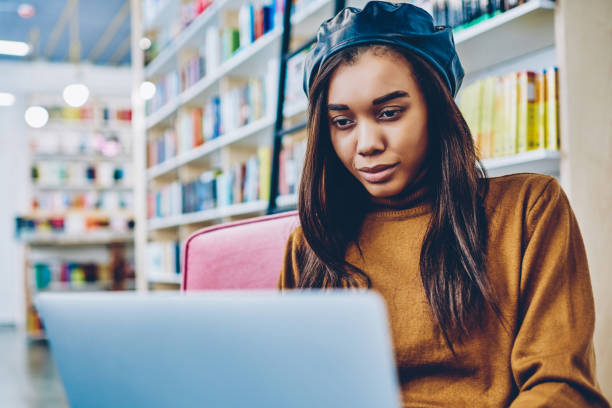 Charming african hipster girl watching webinar on netbook spending time for autodidact in bookstore, young attractive woman reading email from friend and typing answer via modern laptop device