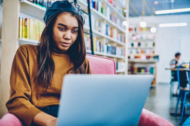 istock Charming african hipster girl watching webinar on netbook spending time for autodidact in bookstore, young attractive woman reading email from friend and typing answer via modern laptop device 1159401604