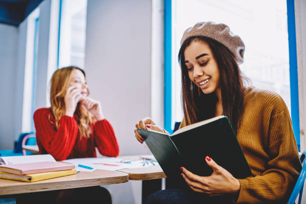 istock Selective focus on happy successful female blogger searching information for next post in positive best seller, cheerful smiling african hipster girl reading book with woman on blurred background 1159401662