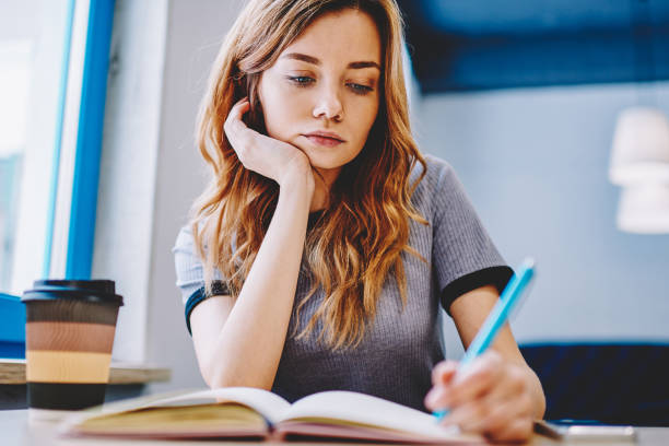 istock Young clever tired female student thinking and writing school homework in textbook for education, thoughtful woman sitting at desktop and spending time for course work indoors notes main theses 1159401942
