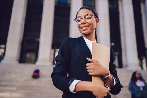 Half length portrait of successful African American jurisconsult dressed in formal wear with folder in hand smiling at camera while standing on courthouse building.Experienced successful female lawyer