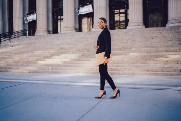 Full length portrait of African American businesswoman with promising career dressed in elegant suit looking at camera while walking near courthouse building. Copy space area for financial information retrato de cuerpo entero de una mujer de negocios afroamericana con una carrera prometedora vestida con un traje elegante mirando la cámara mientras camina baña cerca del edificio del tribunal. copiar área de espacio para obtener información financiera