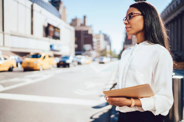 istock Side view of charming african american student of law faculty holding folder with documents in hands standing in downtown in urban setting.Pondering successful dark skinned employee in urban setting 1159407355
