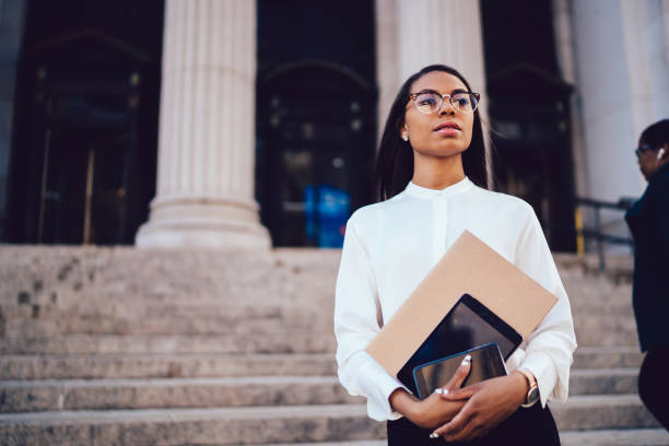 istock Confident dark skinned student of faculty of law looking staight while holding modern smartphone and touch pad in hands strolling near university building.Serious female office worker with folder 1159407404