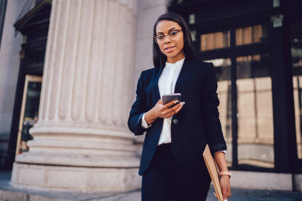Half length portrait of female jurisconsult dressed in black corporate clothing received using cellphone.Experienced entrepreneur lawyer in formal wear holding smartphone standing near office building retrato de media longitud de jurisconsulto femenino vestido con ropa corporativa negra recibido usando el teléfono celular. abogado emprendedor experimentado en ropa formal sosteniendo teléfono inteligente de pie cerca de la construcción de oficinas