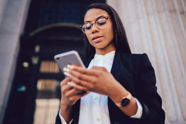 Below view of African american entrepreneur dressed in elegant suit making online payment via banking service on smartphone device standing outdoors. Businesswoman using application on cellphone a continuación, la vista del empresario afroamericano vestido con traje elegante haciendo pago en línea a través del servicio bancario en el dispositivo de teléfono inteligente de pie al aire libre. mujer de negocios usando la aplicación en el teléfo