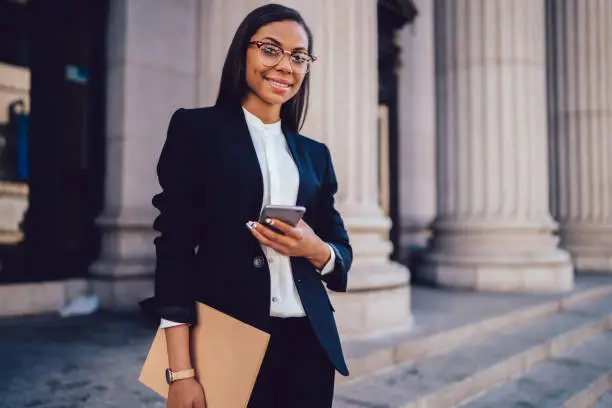 Photo of Portrait of successful African American businesswoman dressed in stylish suit holding in hand folder and mobile phone while standing outdoors near financial office, young woman lawyer using smartphone