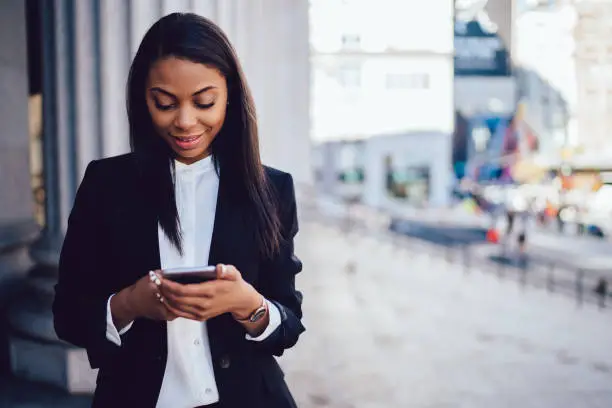 Photo of Positive african american businesswoman dressed in formal wear laughing during messaging online on smartphone device via 4G internet.Cheerful dark skinned female office worker installing application