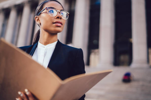 femme d'affaires afro-américaine réfléchie dans des lunettes optiques retenant des documents regardant loin tout en restant dans le cadre urbain avec l'espace de copie. étudiant à la peau foncée femelle de l'université économique élevée - institutional photos et images de collection